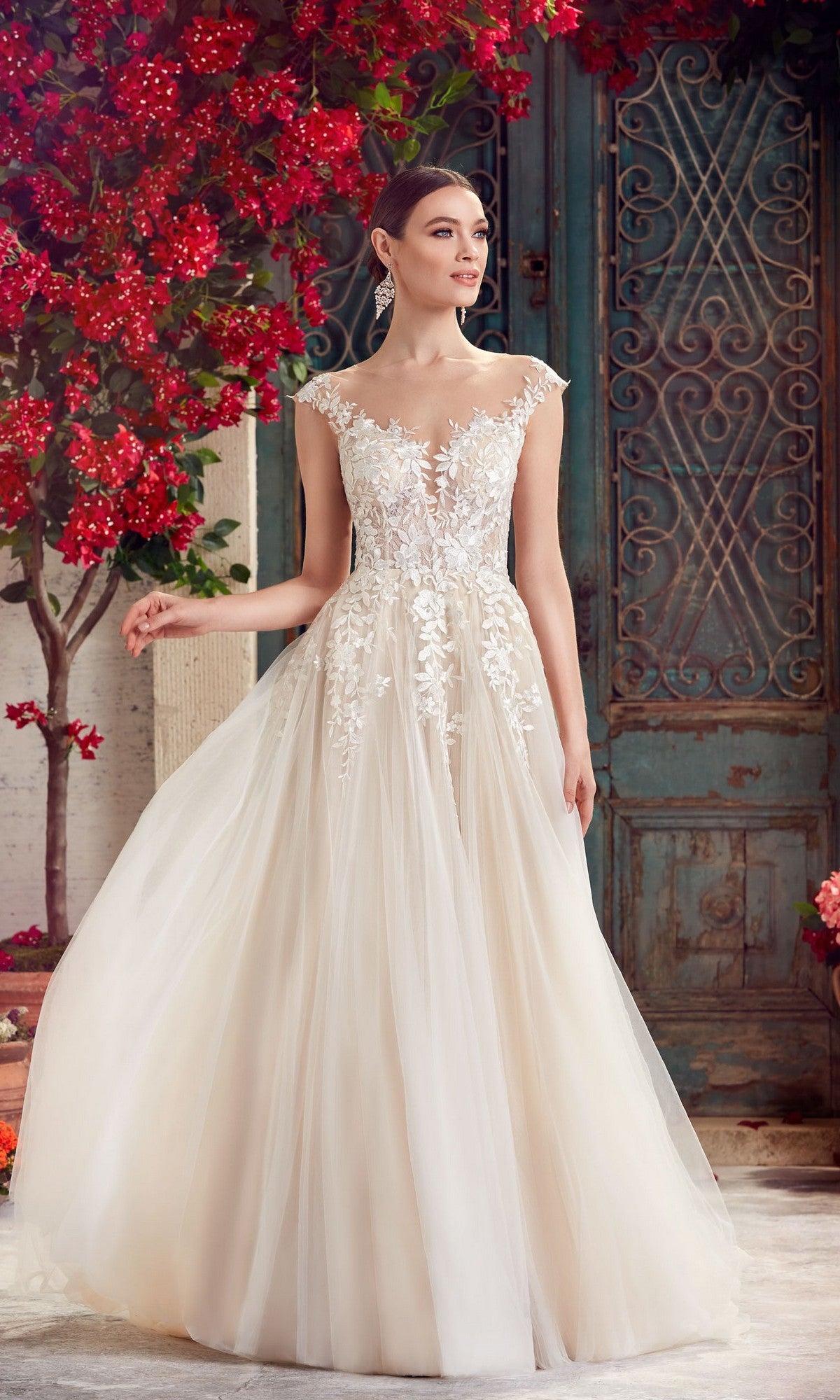 A woman in the Alyce 5090 long formal dress in ivory/champagne, featuring lace embroidery and an a-line tulle skirt, stands near a vintage blue door with red flowering plants.